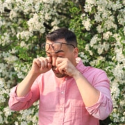 man in pink button up shirt rubbing his eyes in front of a blooming tree